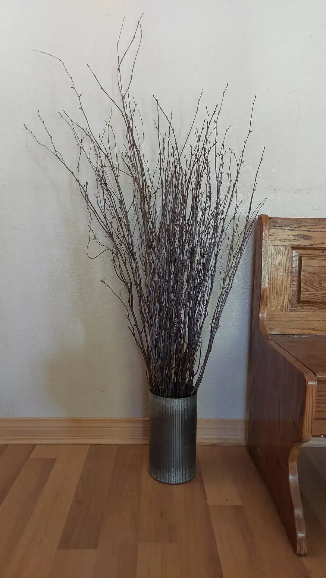 Dried twig arrangement in metal vase on wooden floor.